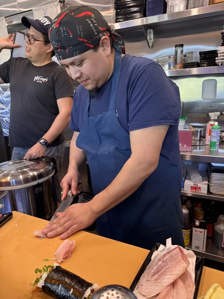 The sushi chef slicing fish on a wooden board while a teammate talks behind him.