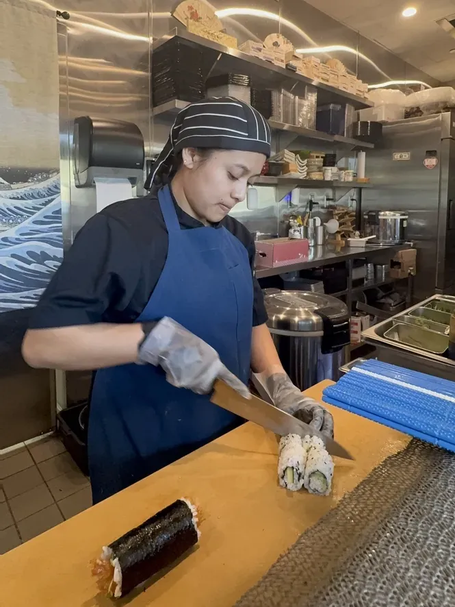A sushi cook cutting a roll in front of a wave mural.